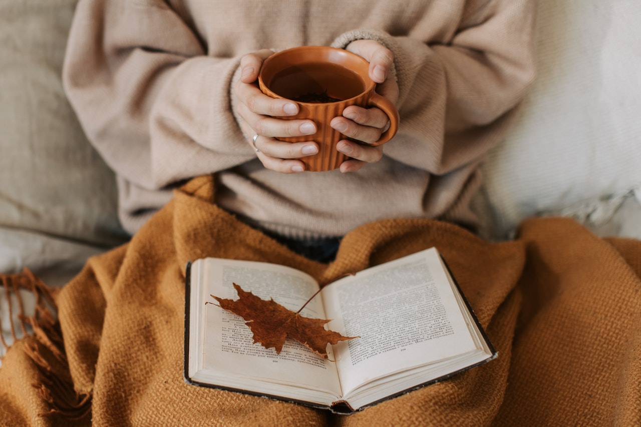 wat je kunt doen tegen de herfstdip is mild zijn voor jezelf, foto van jonge vrouw met dekentje en thee op de bank